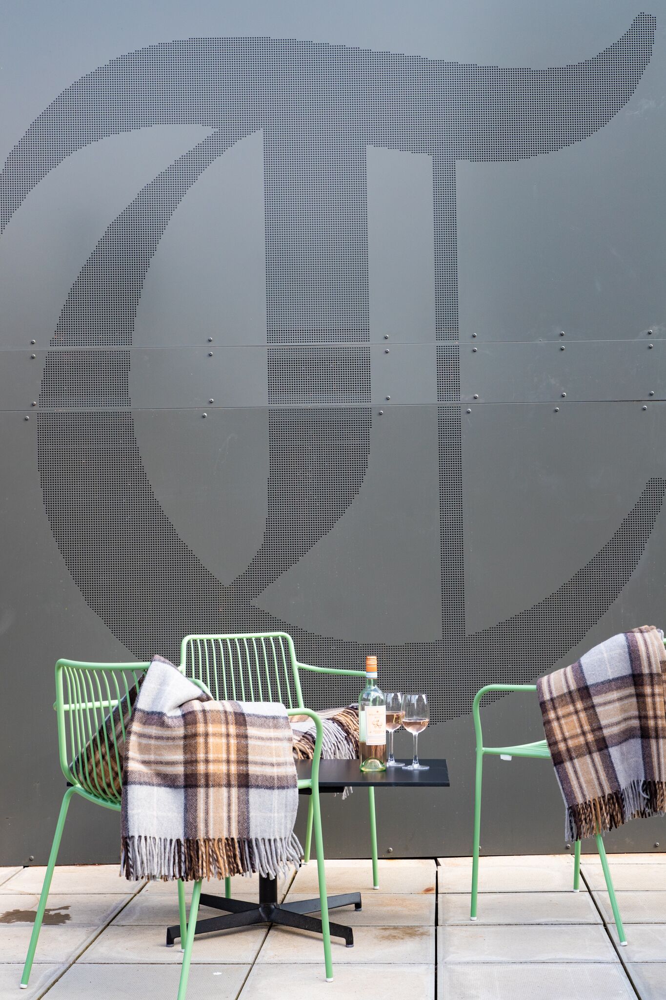 A table with a bottle and glasses, two chairs with blankets, and a large wall mural.