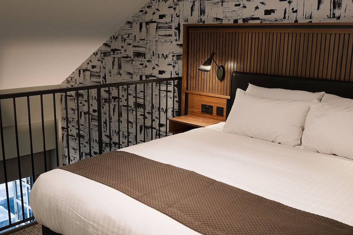 A hotel room with a large bed, a wooden headboard, a nightstand, a wall-mounted lamp, and a balcony with a railing.