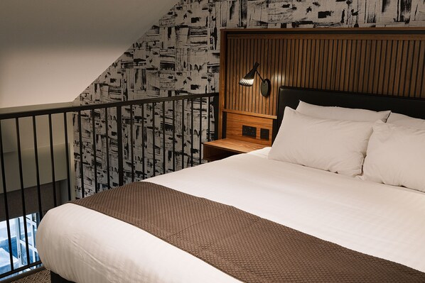 A hotel room with a large bed, a wooden headboard, a nightstand, a wall-mounted lamp, and a balcony with a railing. - Telegraph Hotel - Coventry (Coventry)
