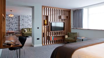 A hotel room with a large bed, a TV, a wooden entertainment unit, a dining area with a table and chairs, and a patterned wallpaper.