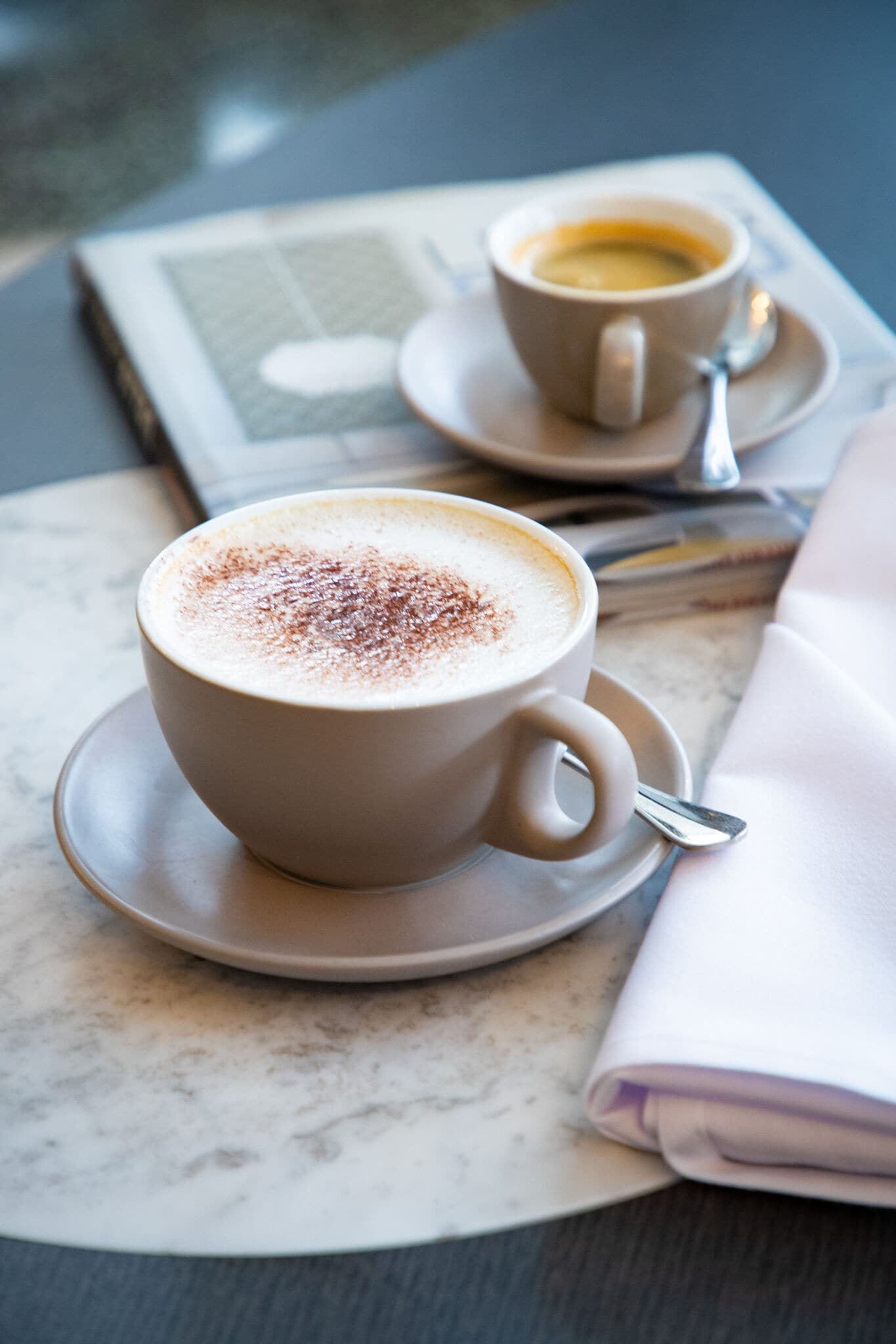 A cup of coffee with a frothy top on a saucer, placed on a table with a newspaper and a folded napkin.