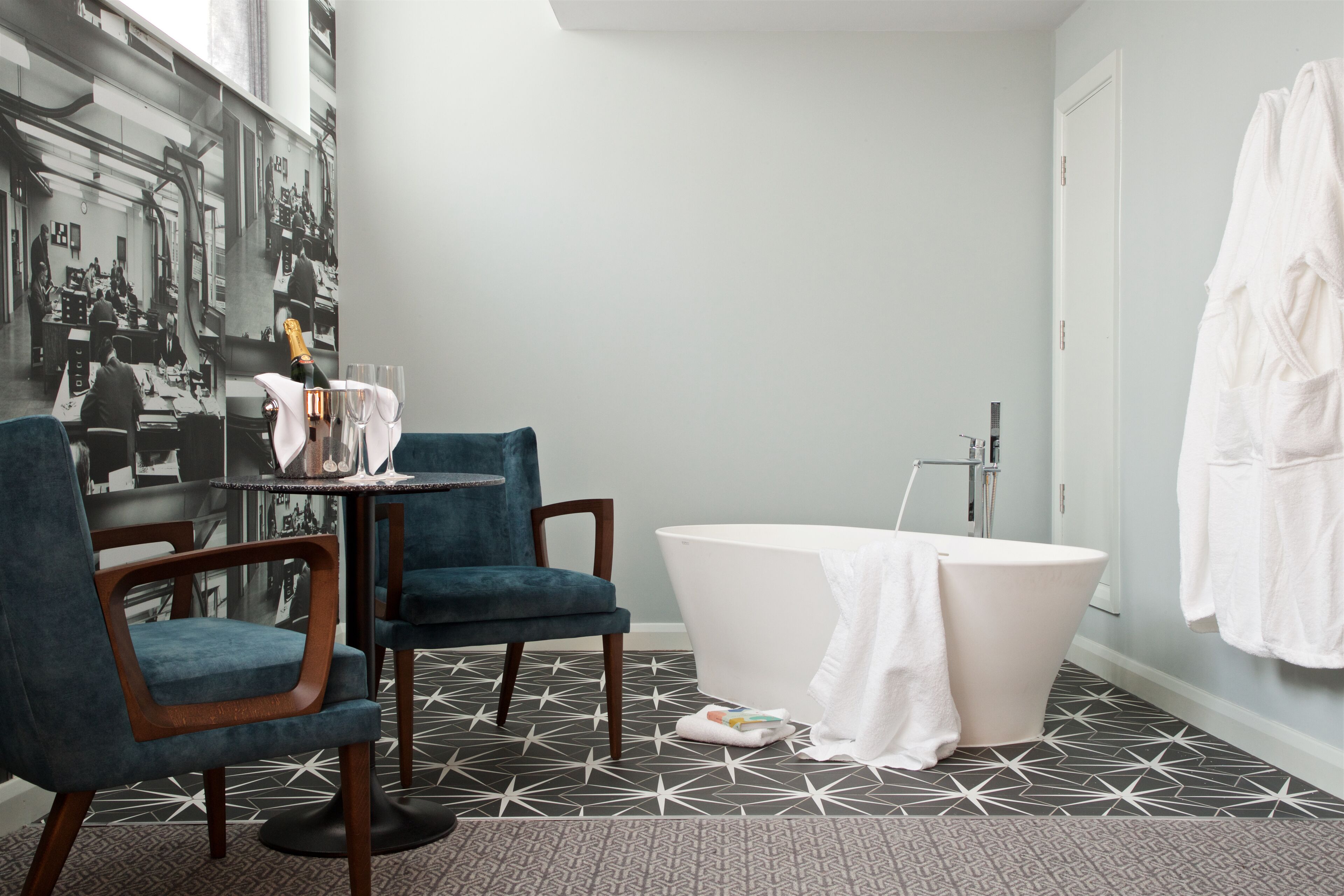 A bathroom with a bathtub, two chairs, a table with a bottle and glasses, and a towel on the floor.