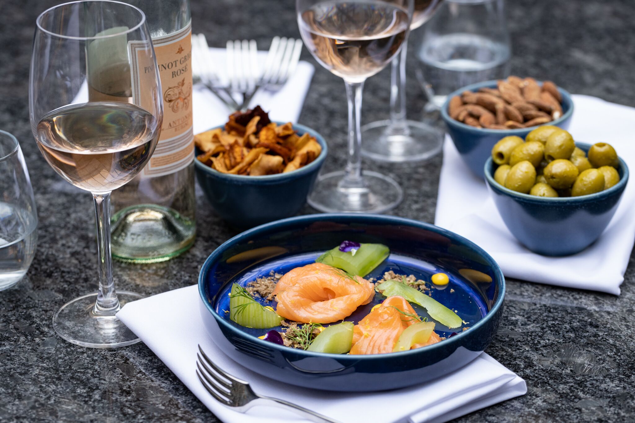 A dining table with a blue bowl of food, wine glasses, and bowls of nuts and olives.