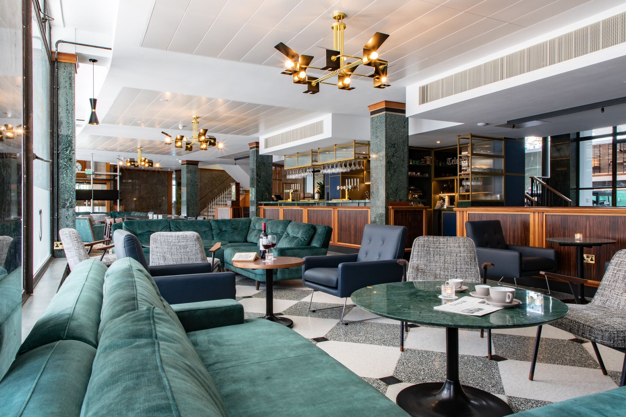 A modern lounge area with green upholstered seating, a bar with a mirrored backsplash, and a staircase leading to an upper level.