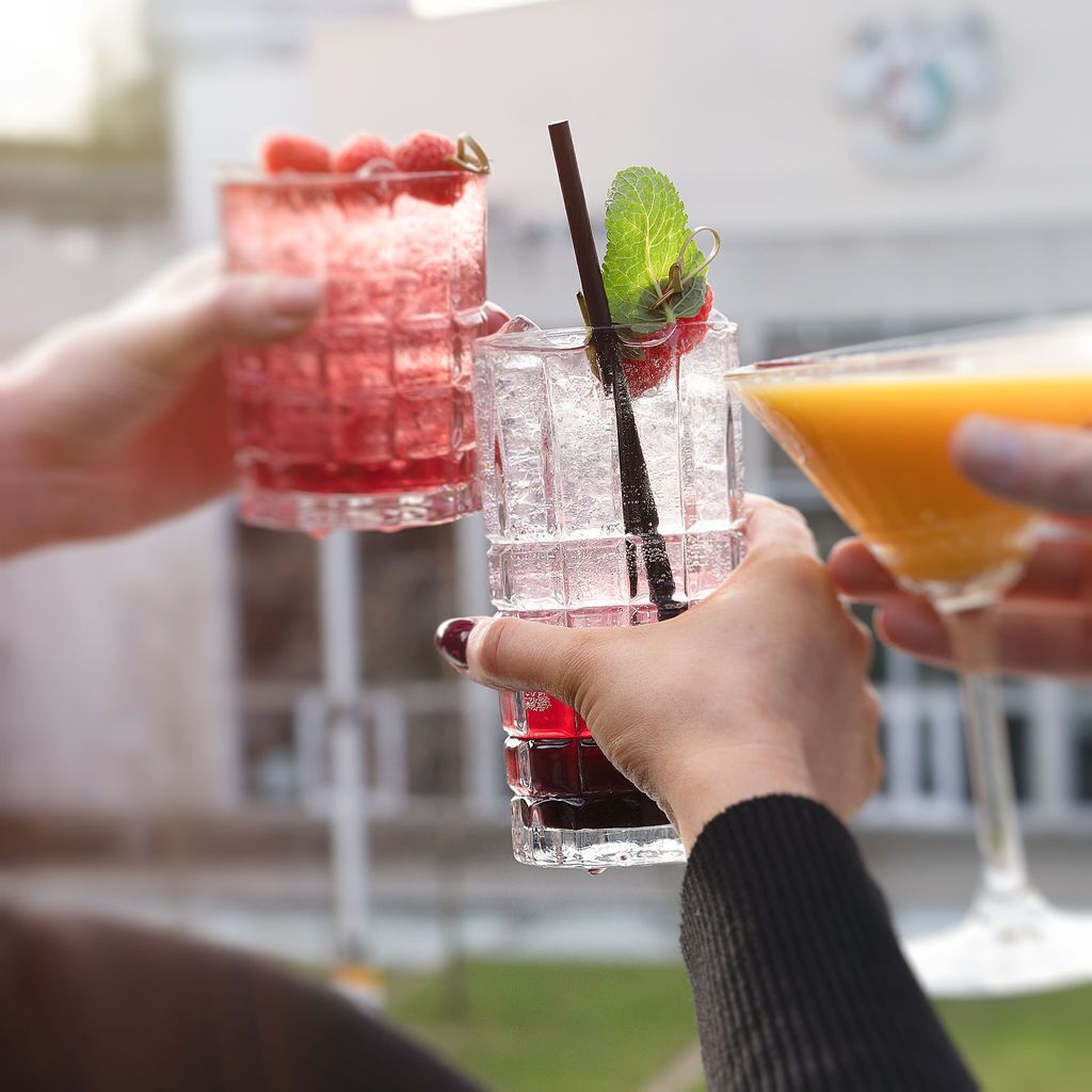 Two people holding glasses of drinks, one with a red beverage and the other with an orange beverage, in a setting with a white building in the background.