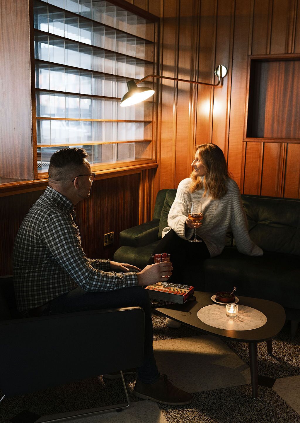 A man and a woman sitting on a green couch in a warmly lit room with wooden paneling, a table with a candle, and a book.