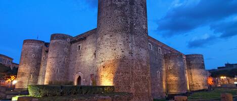 Un castillo de piedra histórico con múltiples torres y una gran torre central, rodeado de un jardín bien cuidado y un paseo pavimentado.