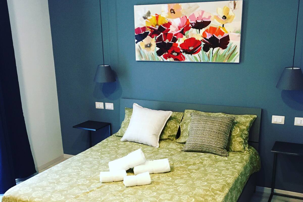 Una cama con una colcha verde estampada, almohadas blancas y toallas enrolladas. En la pared sobre la cama hay un cuadro con flores.