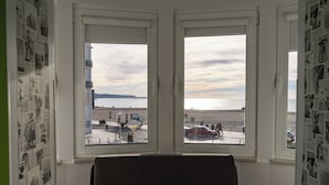 A room with a large window overlooking a beach, a chair in the foreground, and walls decorated with black and white photos. - By The Sea  (Nazaré)