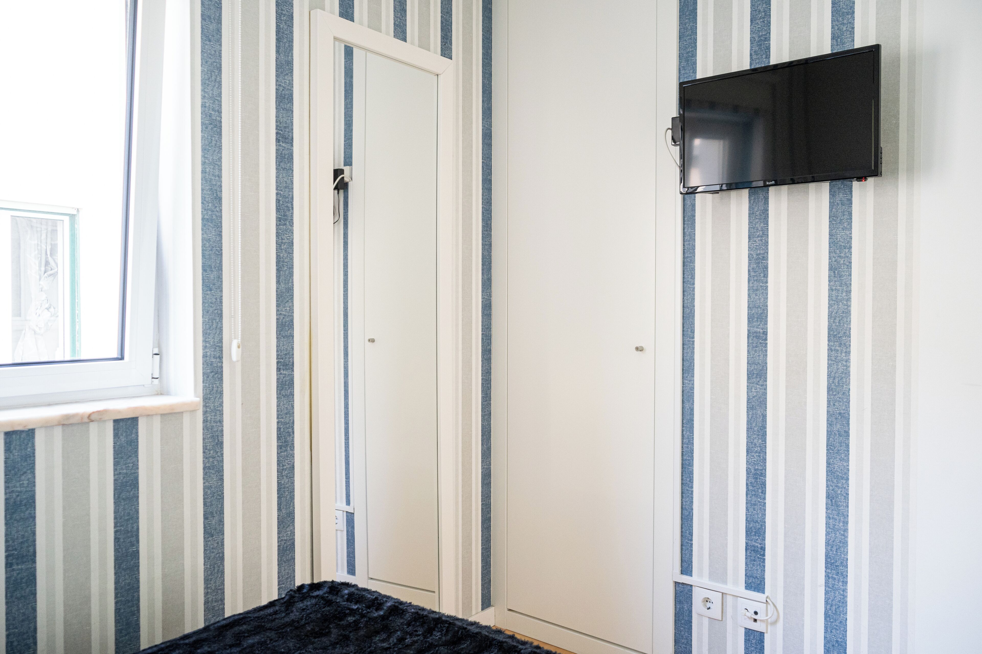 A room with striped wallpaper, a closed door, a mounted TV, and a window.