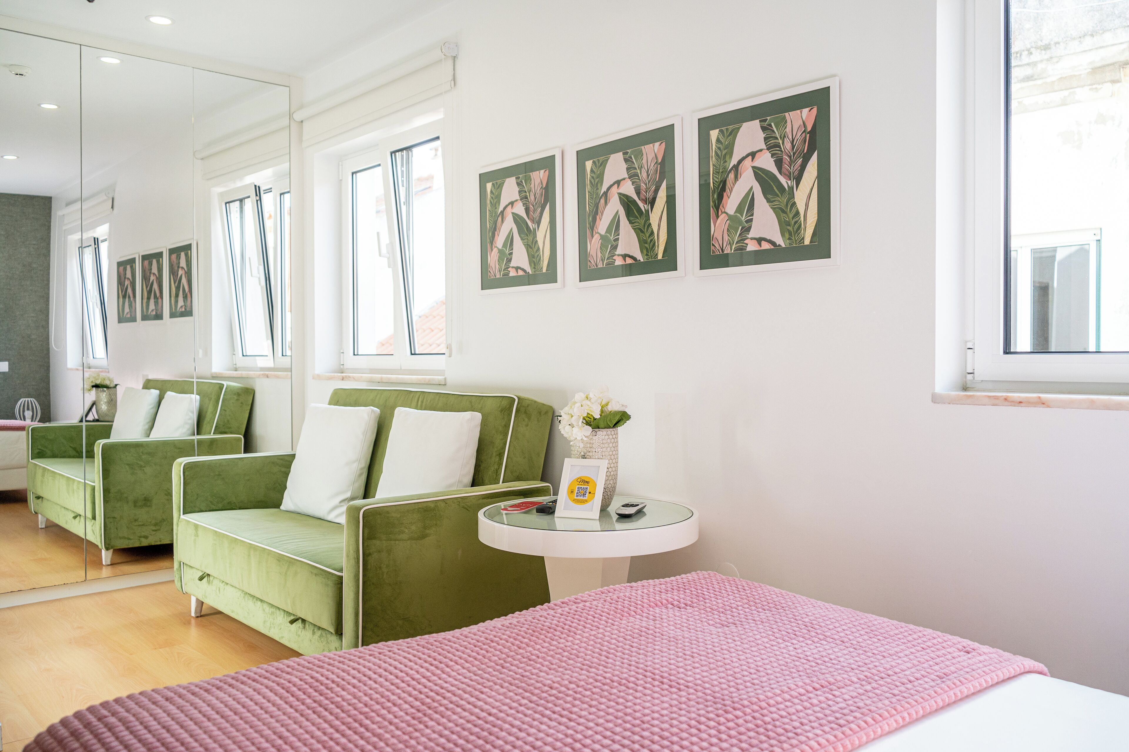 A modern hotel room with a green sofa set, a pink bedspread, and framed botanical prints on the wall.