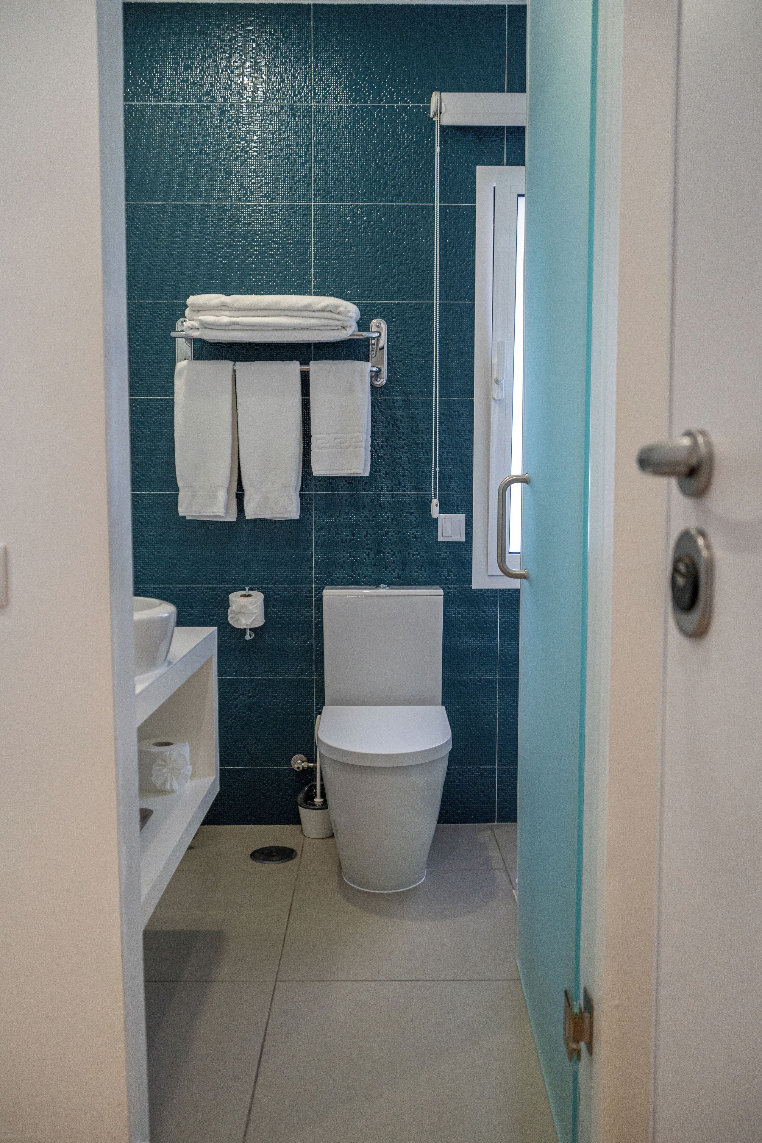 Une salle de bain avec des toilettes blanches, un lavabo et une étagère contenant des articles de toilette. Des serviettes sont suspendues à un porte-serviettes et il y a une fenêtre au-dessus des toilettes.