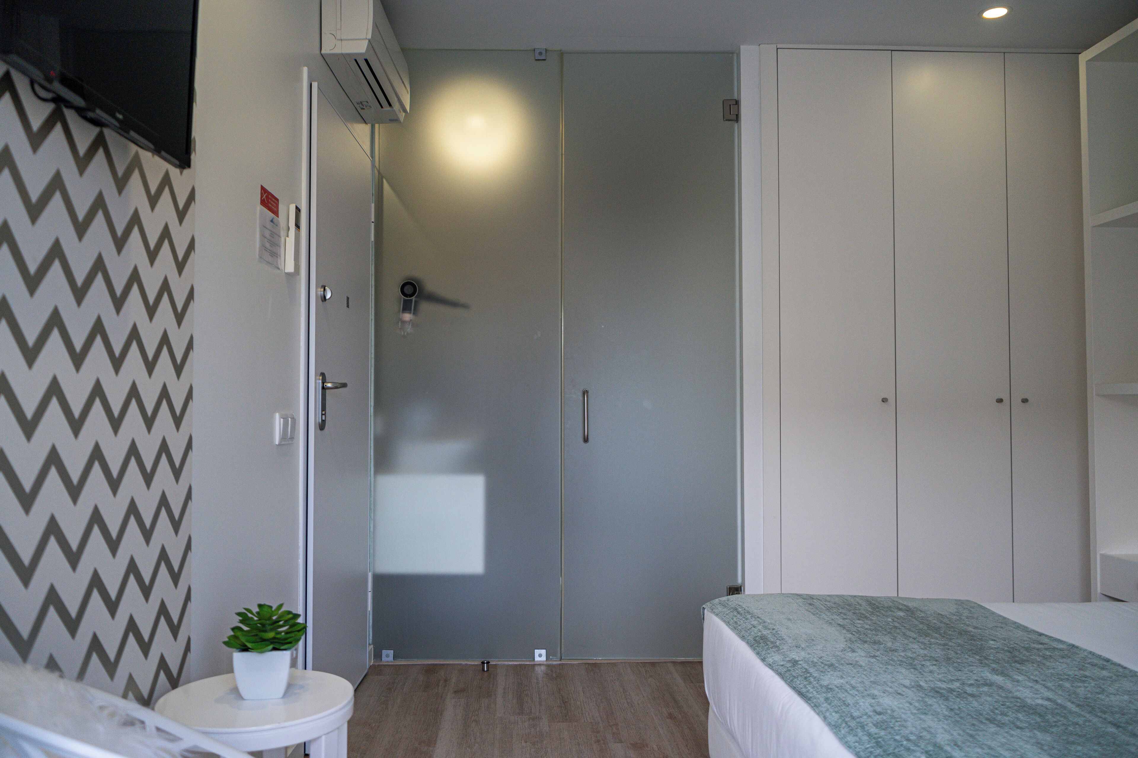 A bedroom with a bed, a small table with a plant, a wardrobe, and a door leading to a bathroom.