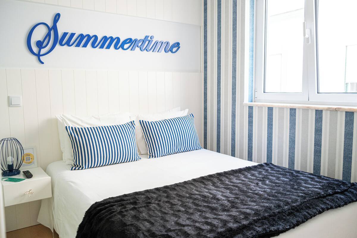 A neatly made bed with striped pillows, a black throw blanket, and a wall sign that reads "Summertime".