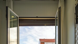 A room with a bed, a chair, and a large window offering a view of buildings. - By The Sea  (Nazaré)