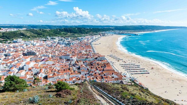 A coastal town with a beach, red-roofed buildings, and a clear blue sea. - By The Sea  (Nazaré)