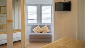 A room with a bed, a grey sofa with cushions, a television on the wall, and a window with a view. - By The Sea  (Nazaré)
