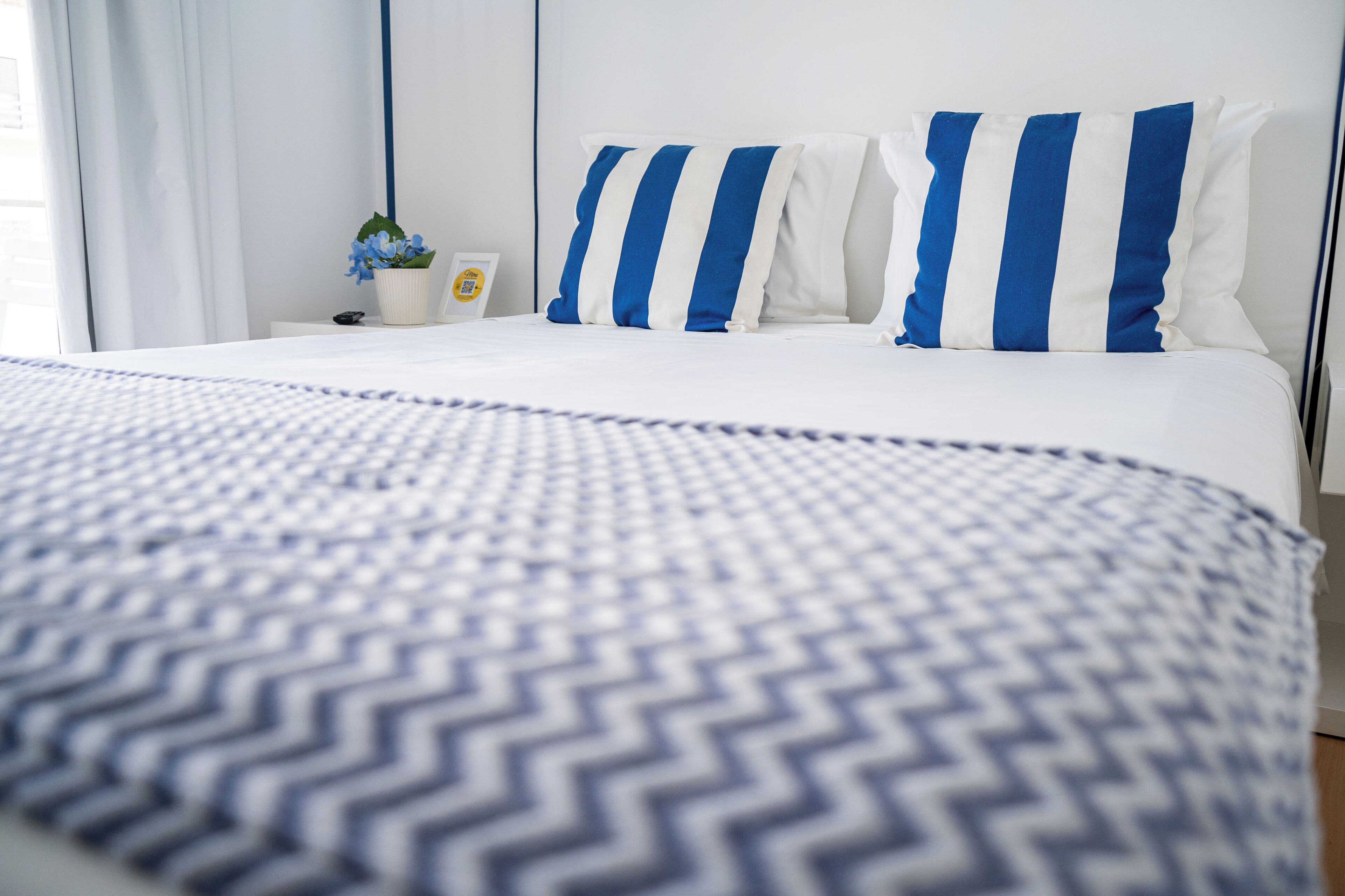 A neatly made bed with a blue and white striped pillow, a small potted plant, and a framed picture on a bedside table.