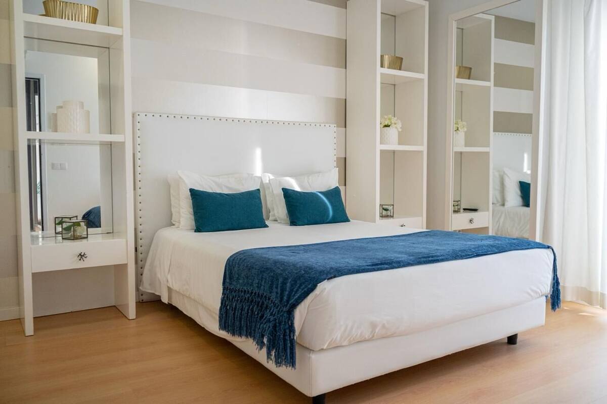 A bedroom with a large bed, white headboard, and blue bedding. There are white shelves with various items on them.