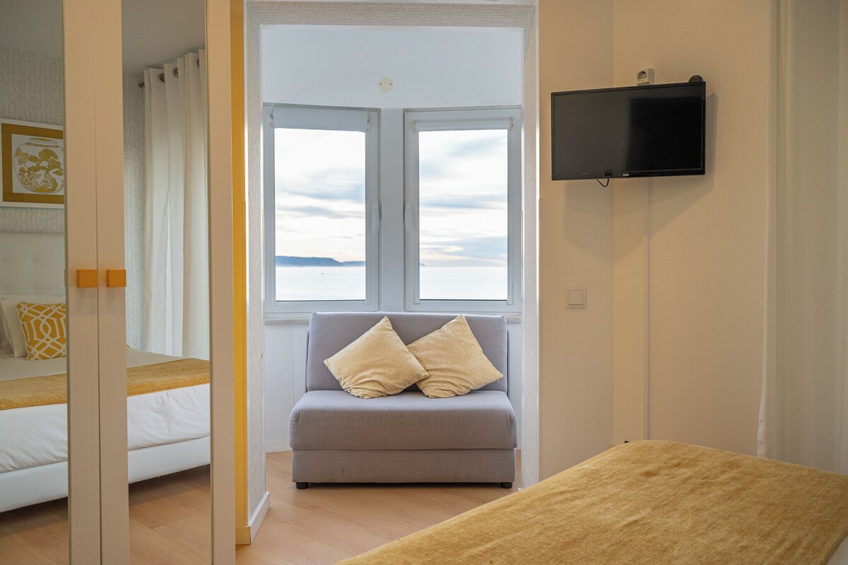 A room with a bed, a grey sofa with cushions, a television on the wall, and a window with a view.
