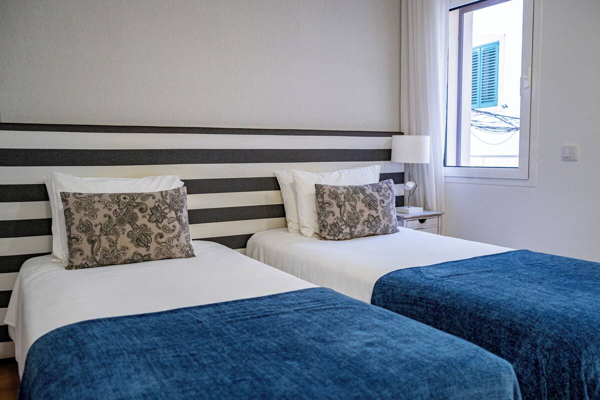 A hotel room with two beds, each with a patterned pillow and a blue blanket.