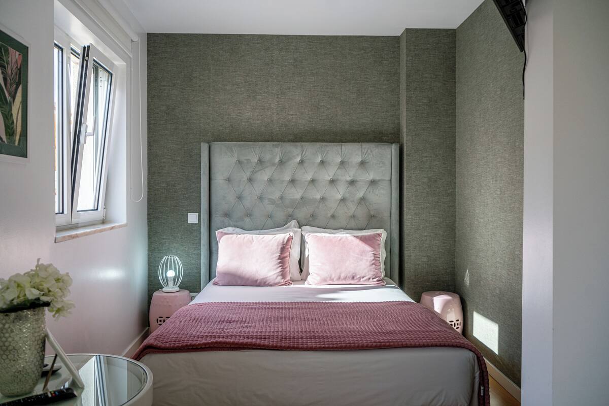 A bedroom with a tufted headboard, two pillows, a pink blanket, a bedside table with a lamp, and a window with curtains.