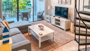 A modern living room with a sofa, coffee table, and TV. There is a balcony with outdoor furniture. - BT 3322 Fountain/Pond View Condo-Paradise Vacation Rental in Ocean Village (Fort Pierce)