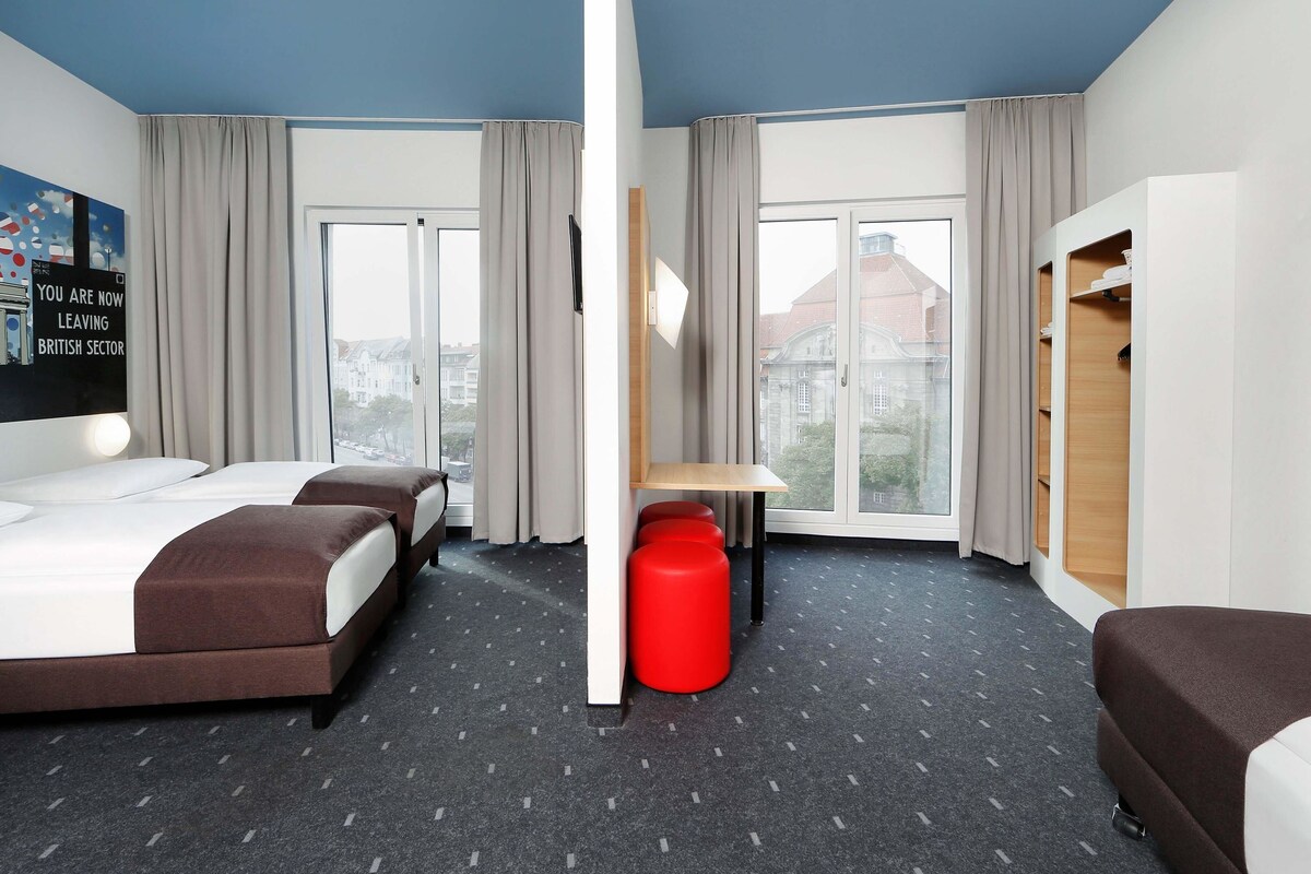 Una habitación de hotel con dos camas, un escritorio, un reposapiés rojo y un armario.