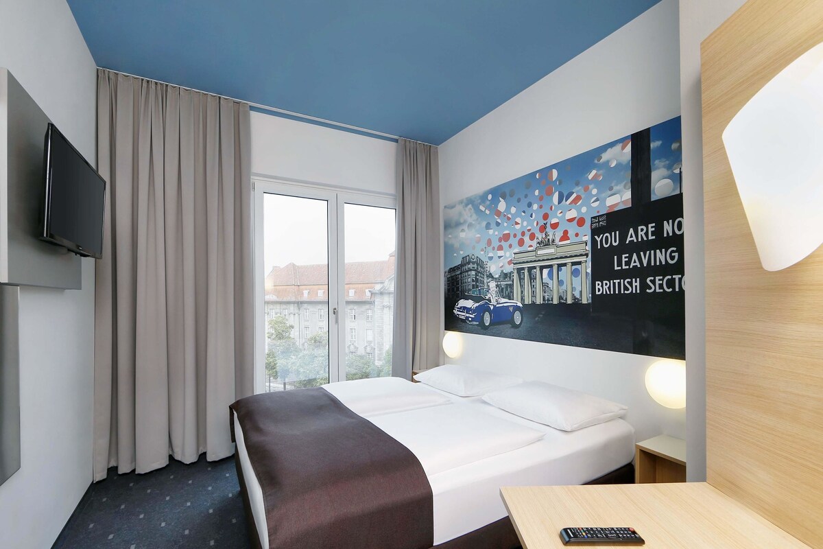 Una habitación de hotel con cama, televisor, ventana con cortinas, pared con un mural de paisaje urbano y escritorio con control remoto.