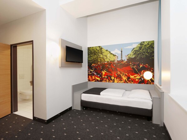 Une chambre d’hôtel avec un lit, un téléviseur à écran plat fixé au mur et une grande fresque représentant une foule devant un monument.