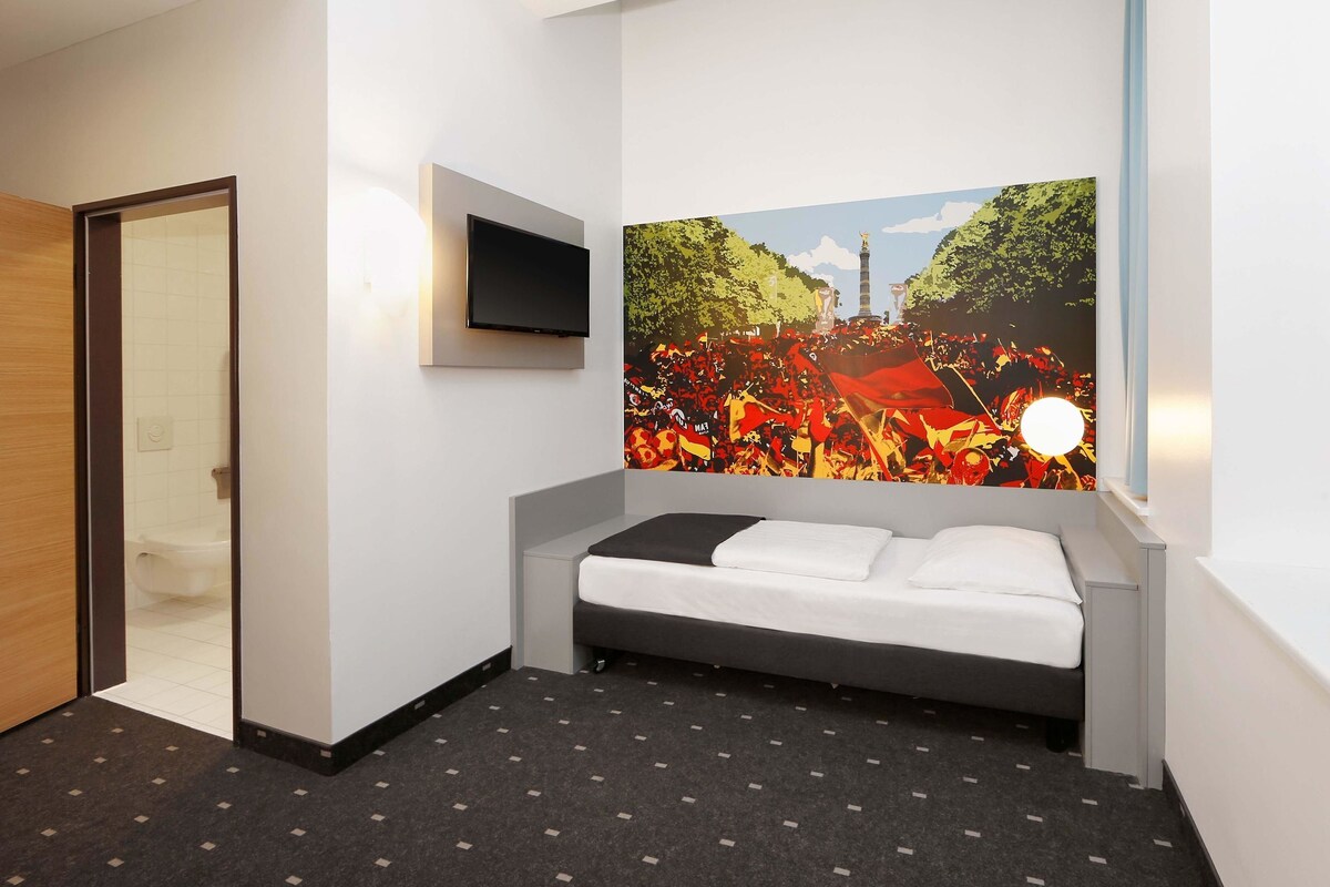 Una habitación de hotel con cama, televisor de pantalla plana montado en la pared y un mural grande que muestra a una multitud junto a un monumento.