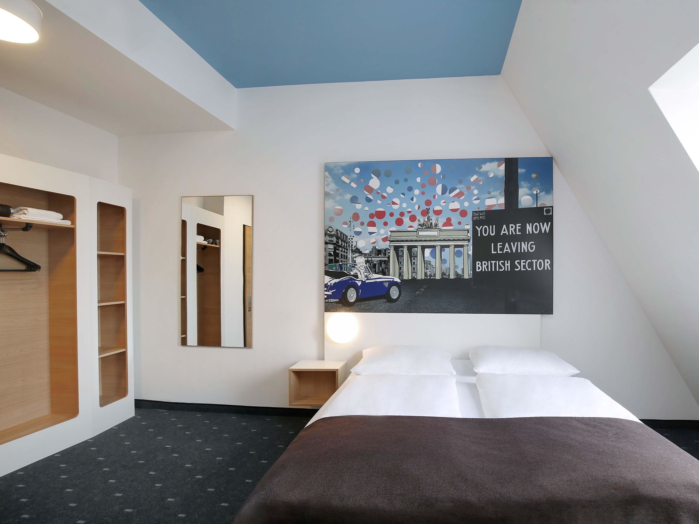Quarto de hotel com uma cama, um guarda-roupa, um espelho e uma parede com uma pintura da paisagem urbana.