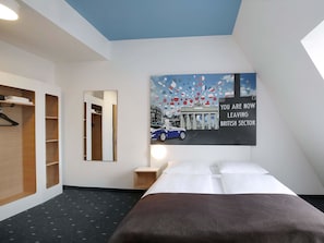 Une chambre d’hôtel avec un lit, une armoire, un miroir et un mur orné d’un tableau représentant un paysage urbain.