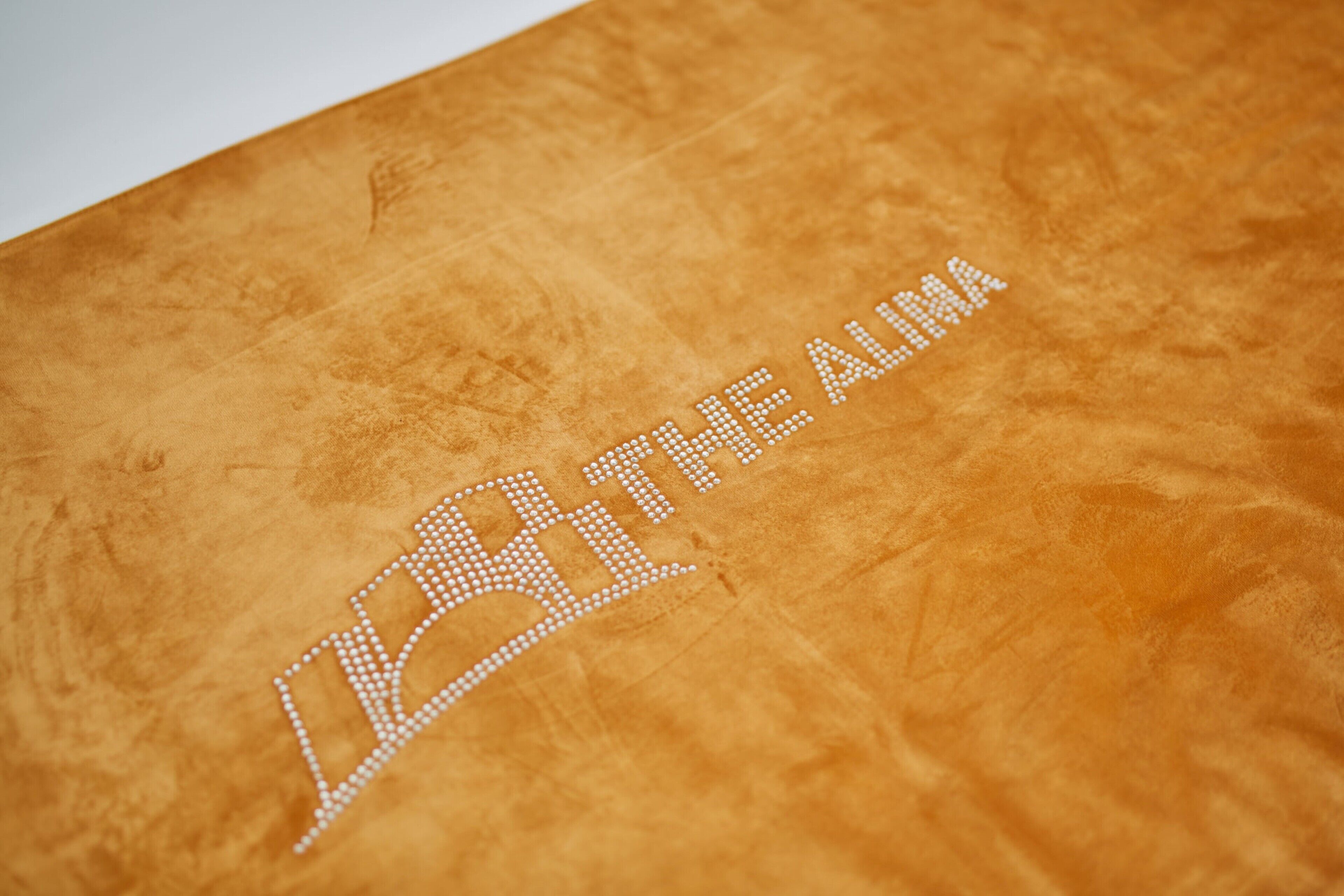 Eine strukturierte, orangefarbene Oberfläche mit einer weißen Stickerei, die "BE THE ALMAH" ausschreibt.
