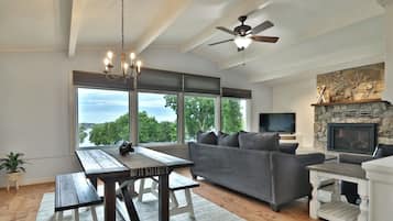 A spacious living room with a grey sofa, a wooden dining table, a ceiling fan, and a stone fireplace.
