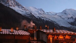 A mountain landscape with snow-covered peaks and a building with a wooden facade and a chimney emitting smoke.