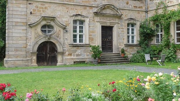 A historic stone building with a central entrance, a small garden with flowers, and a paved walkway leading to the entrance. - Ferienzimmer im Schloss Burgpreppach (Burgpreppach)