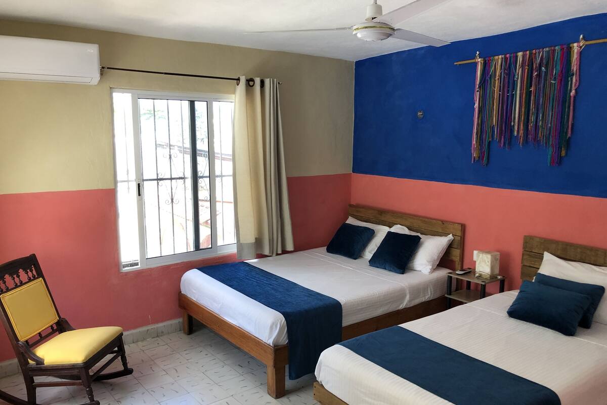 A hotel room with two beds, a rocking chair, a window with curtains, a ceiling fan, and a colorful wall hanging.