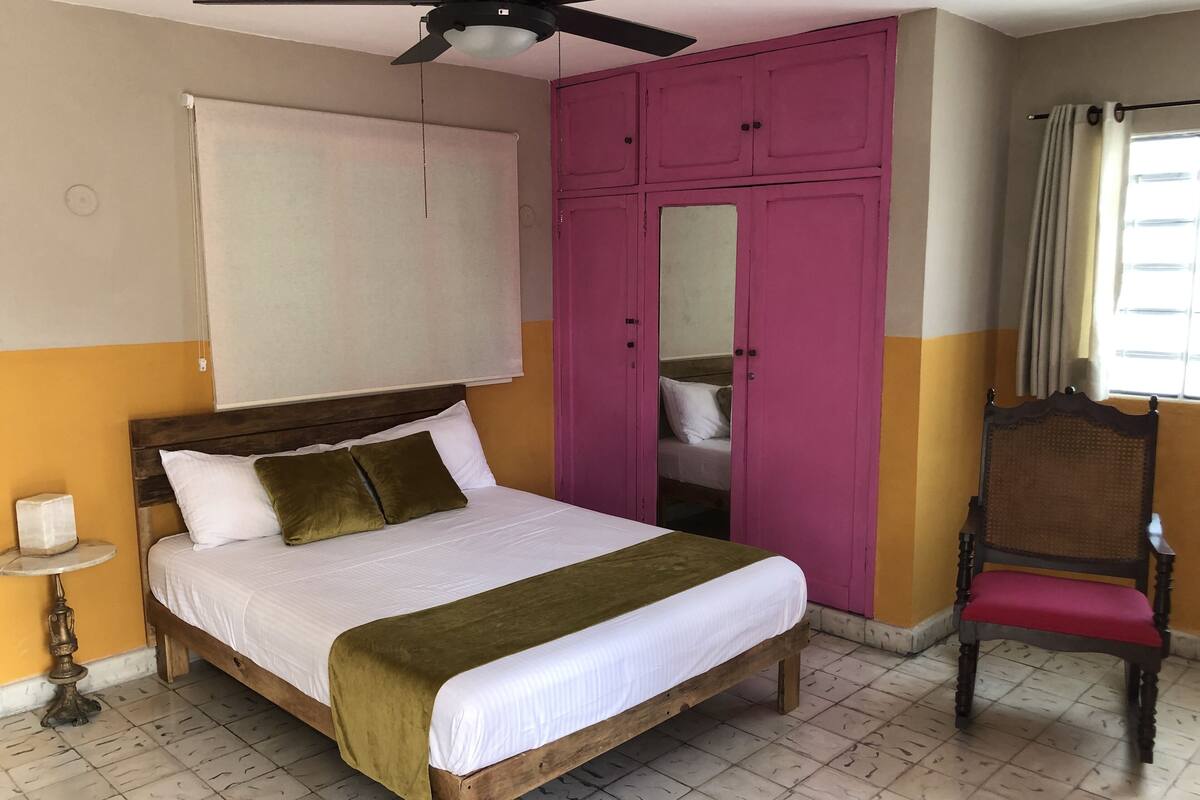 A bedroom with a wooden bed, a ceiling fan, a pink wardrobe, and a chair.