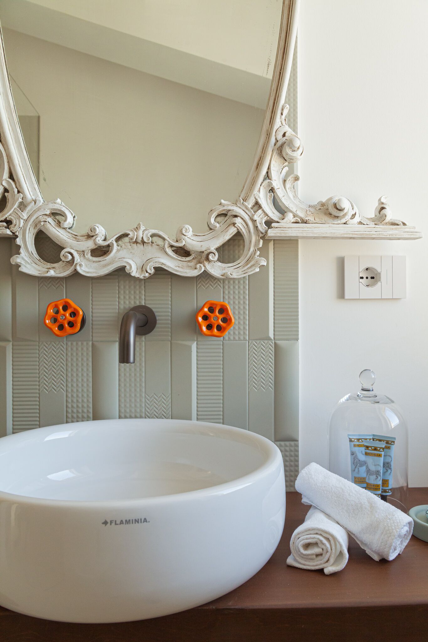 Un baño con lavamanos blanco, espejo decorativo y mandos de ducha anaranjados.