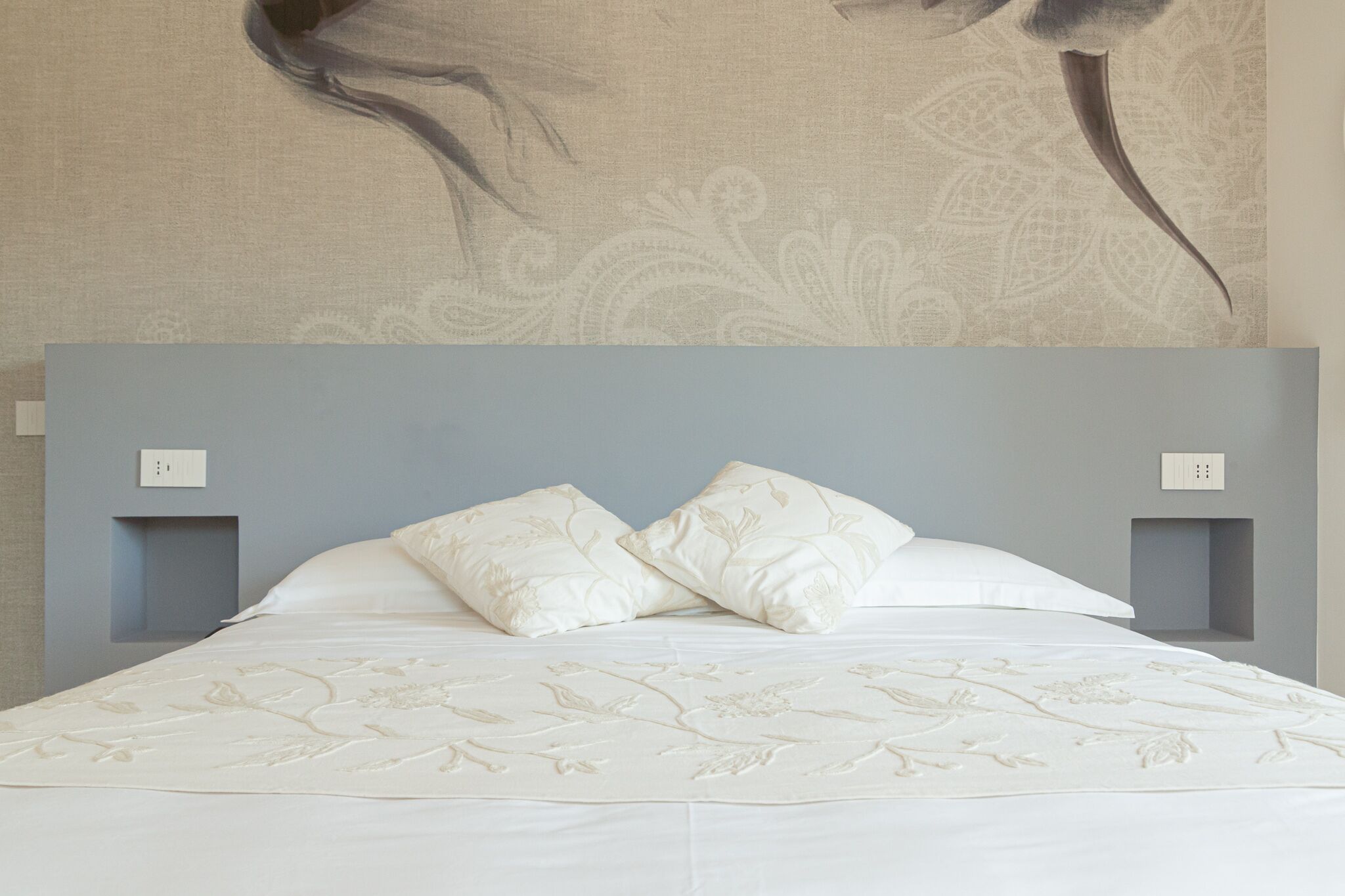A bed with a floral-patterned bedspread and two pillows, a wall with a large abstract painting, and two bedside tables with light blue upholstery.