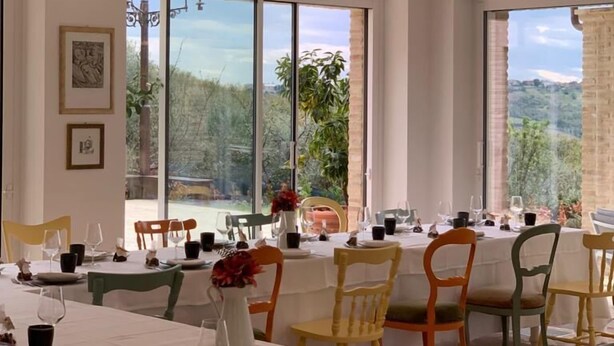 A dining area with a long table set for a meal, large windows with a view of greenery, and hanging pendant lights. - Frantoio L'Olinda (San Marcello)