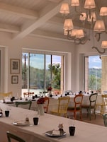 A dining area with a long table set for a meal, large windows with a view of greenery, and hanging pendant lights.