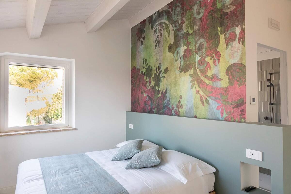 Une chambre avec un lit, une fenêtre, un grand décor mural représentant des motifs floraux et un espace douche.
