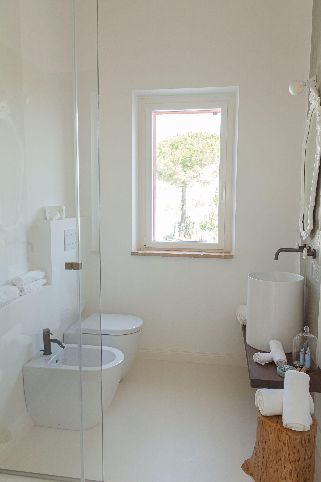 Un baño moderno con cabina de ducha de vidrio, inodoro, bidé y lavamanos. Hay una ventana que proporciona luz natural y vistas a la vegetación exterior.