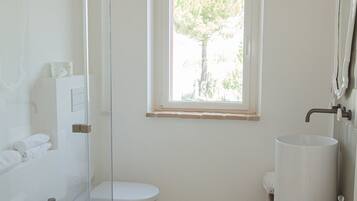 Un baño moderno con cabina de ducha de vidrio, inodoro, bidé y lavamanos. Hay una ventana que proporciona luz natural y vistas a la vegetación exterior.