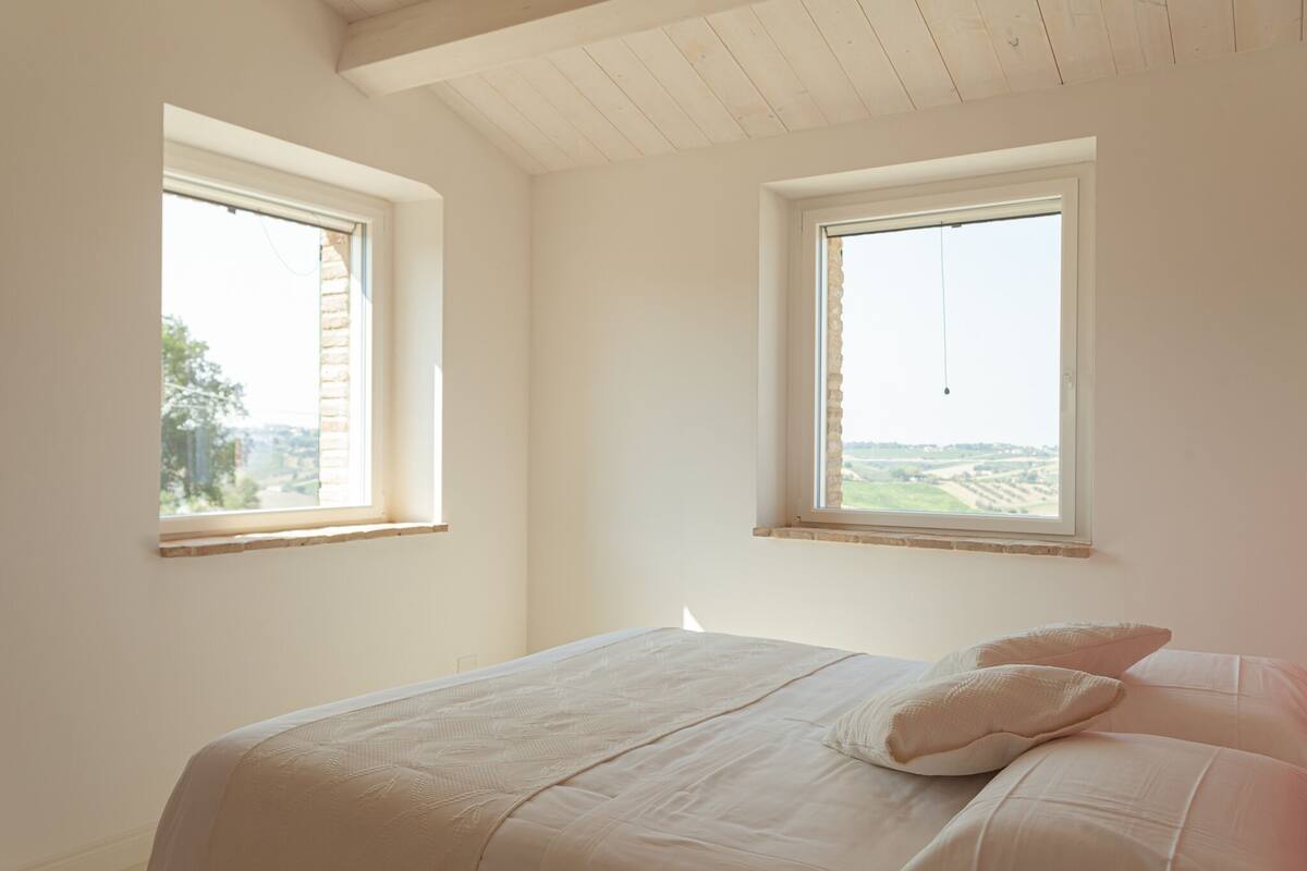 Une chambre avec deux fenêtres, un lit avec des oreillers et une vue sur la campagne.
