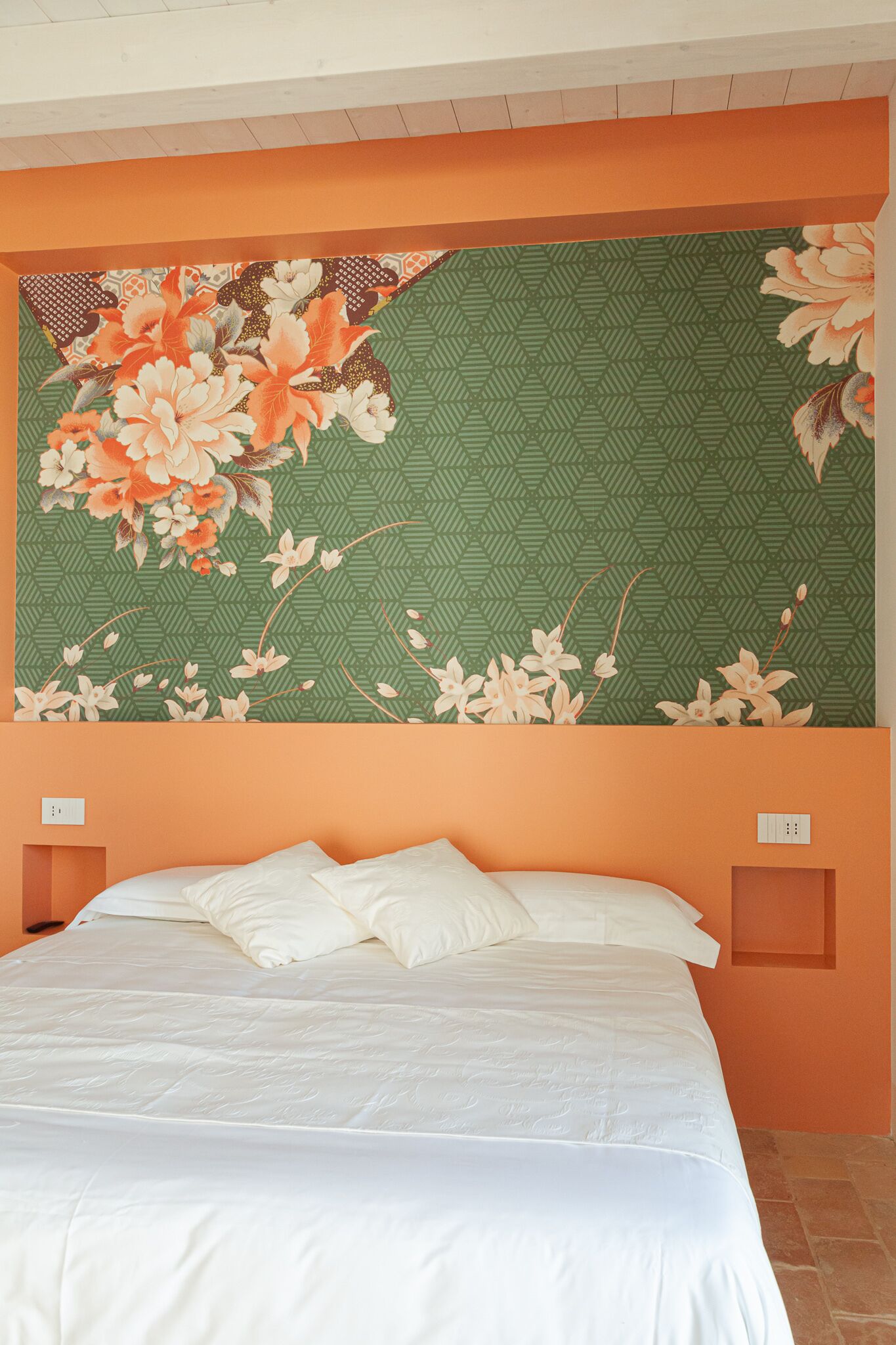 A bed with white bedding in a room with orange walls and a large floral-patterned wall hanging above it.