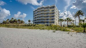 A multi-story hotel with balconies, located on a sandy beach with palm trees.