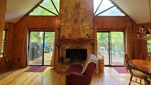A cozy cabin interior with a stone fireplace, wooden walls, and large windows offering a view of the outdoors. - Bear Gone Fishing- Cool Down in the Creek!!! Mins to Highlands (Scaly Mountain)